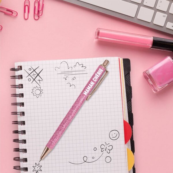 Stylo à bille rétractable avec un corps rose pailleté, des finitions or rose et l'inscription MAMAN CHÉRIE en blanc.