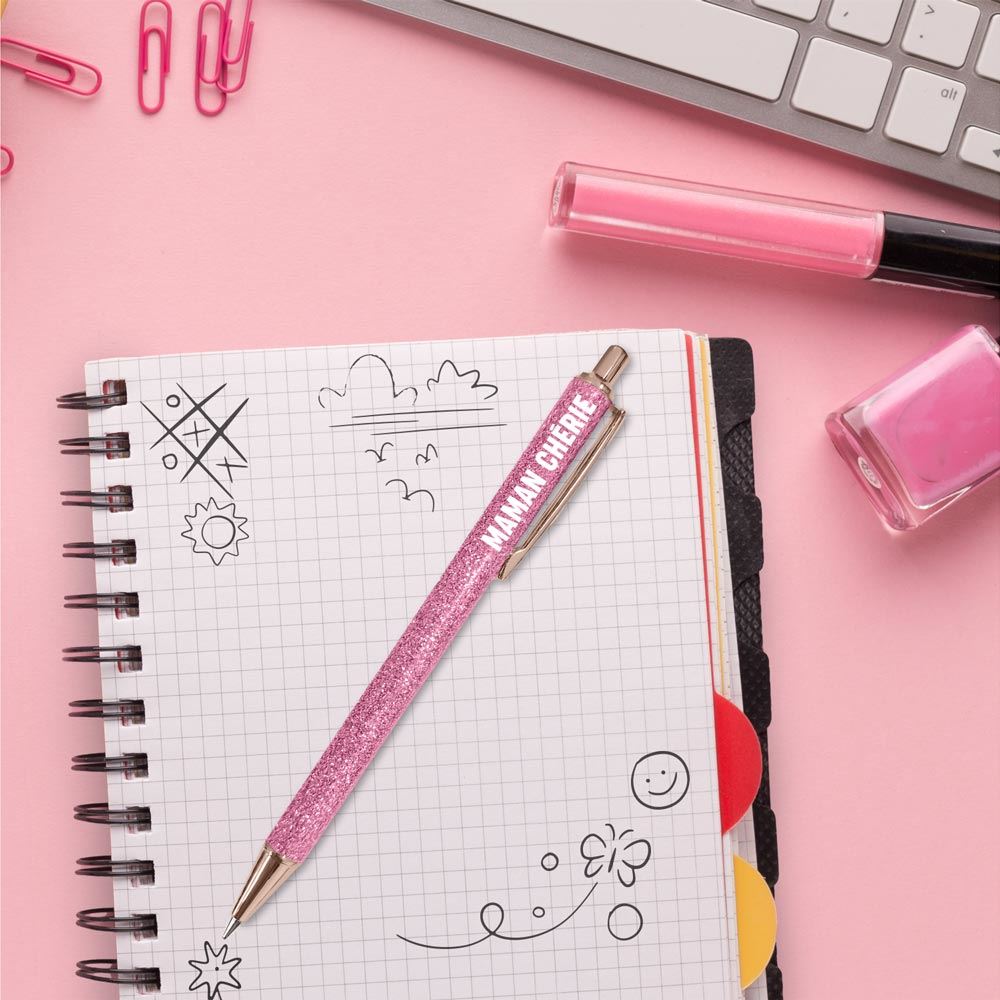 Stylo à bille rétractable avec un corps rose pailleté, des finitions or rose et l'inscription MAMAN CHÉRIE en blanc.