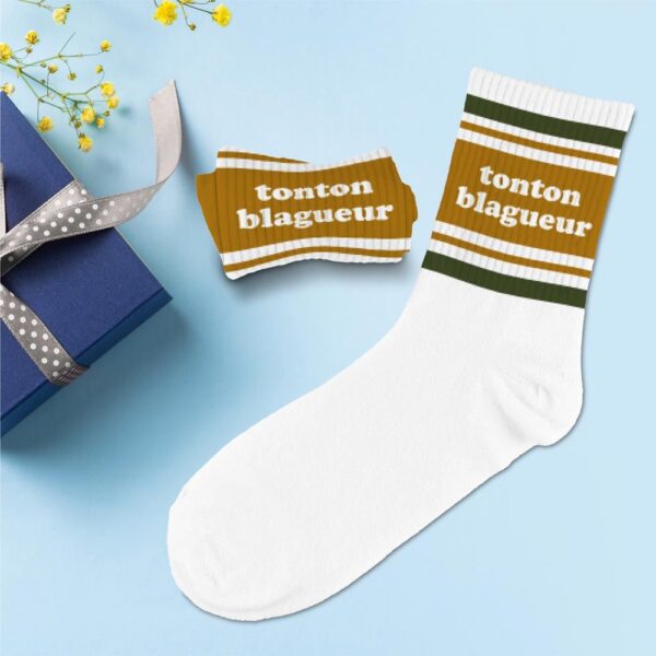 Paire de chaussettes hautes blanches pour homme présentant une large bande ocre avec l'inscription "tonton blagueur" et des fines rayures vertes