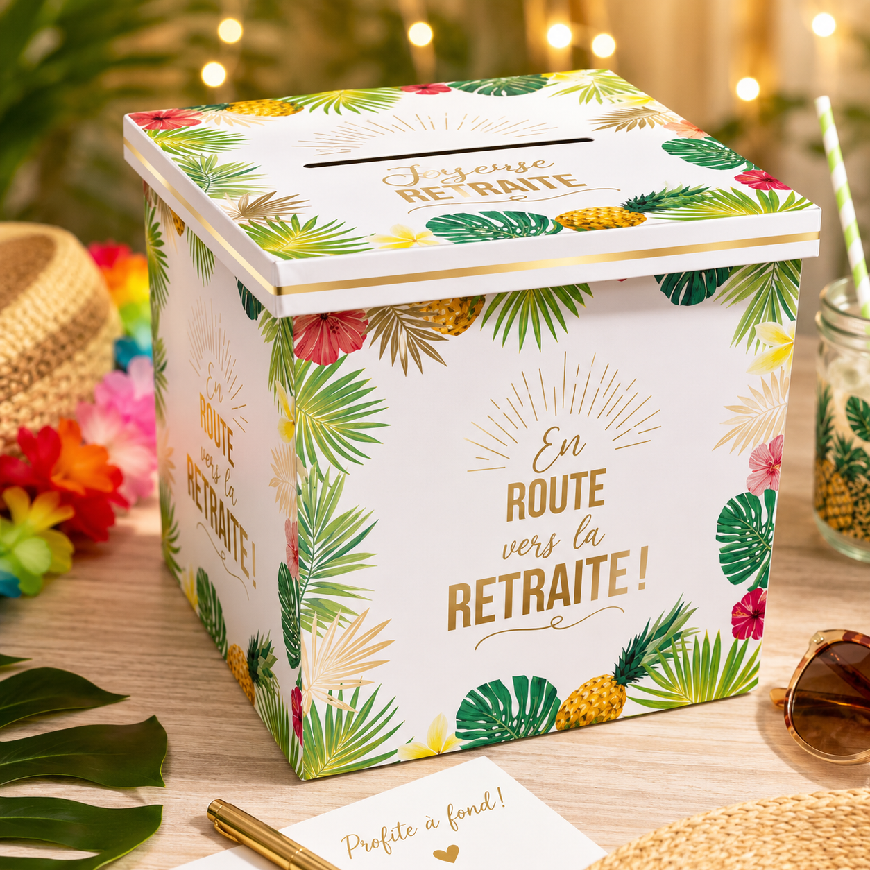 Une urne cubique en carton blanc pour célébrer un départ à la retraite. Elle est décorée de motifs tropicaux colorés : feuilles de palmier vertes, ananas jaunes et fleurs d'hibiscus roses et rouges. Sur les faces, l'inscription "En route vers la RETRAITE !" est écrite en lettres dorées et marron. Le couvercle dispose d'une fente et de l'inscription "Joyeuse RETRAITE"